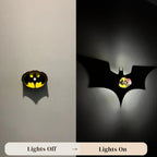 A split comparison showing a Batman logo wall light in two states: on the left, off with a small warm yellow glow inside the bat-shaped outline; on the right, on with a large black bat shadow投射 on a wall and a bright yellow light shining through the center. Text at the bottom reads 'Lights Off' and 'Lights On' with 'www.